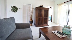 A living room with a grey sofa, a wooden cabinet, a vase with dried plants, a jar with a rope handle, and a remote control on a magazine.