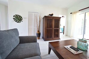 A living room with a grey sofa, a wooden cabinet, a vase with dried plants, a jar with a rope handle, and a remote control on a magazine.