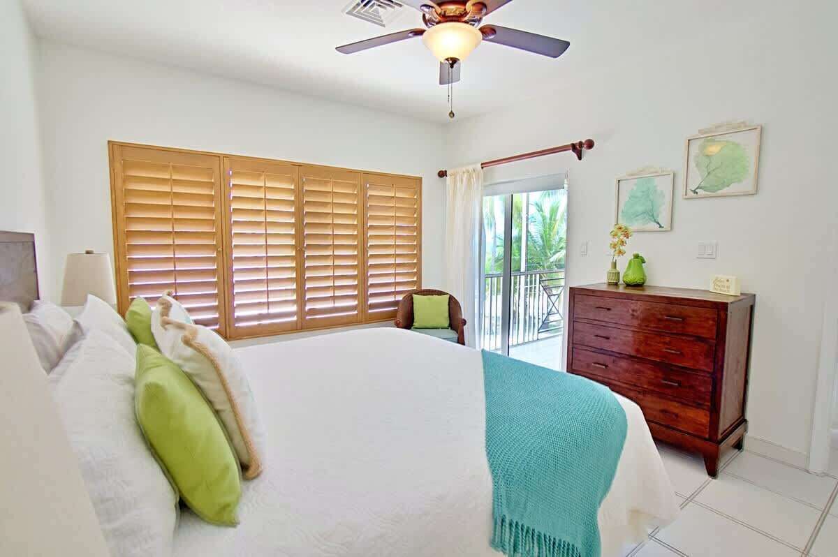 Un dormitorio con cama, persianas de madera, ventilador de techo, cómoda y balcón con vistas a la vegetación.