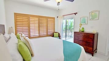 Un dormitorio con cama, persianas de madera, ventilador de techo, cómoda y balcón con vistas a la vegetación.