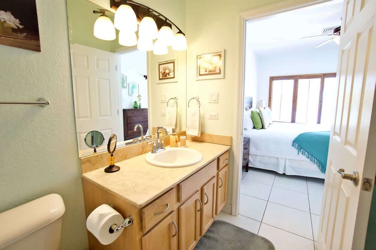 A bathroom with a sink, mirror, and toilet. There is a door leading to a bedroom with a bed and a window.