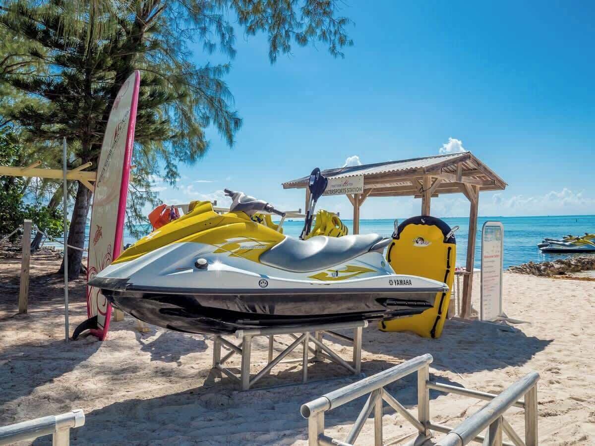 Un jet ski sobre un soporte cerca de una playa, con un pabellón y una tabla de surf al fondo.