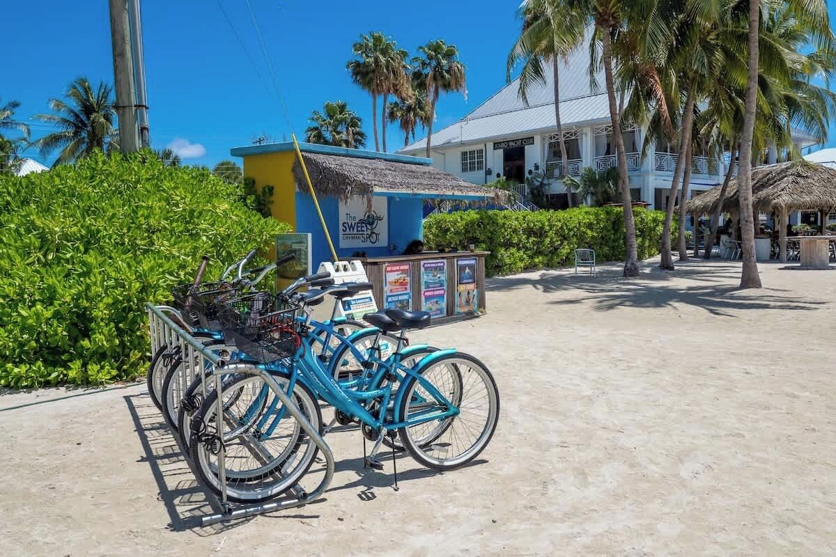 Una estación de alquiler de bicicletas, con una bicicleta azul en primer plano, una estructura con techo de paja y un edificio con balcones al fondo.