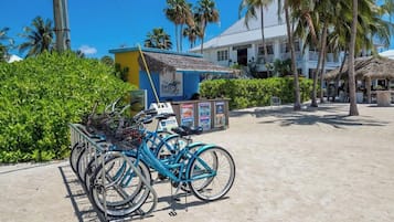 Una estación de alquiler de bicicletas, con una bicicleta azul en primer plano, una estructura con techo de paja y un edificio con balcones al fondo.