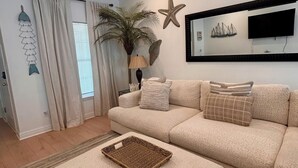 Sala de estar com sofá bege, uma mesinha, decoração com estrela-do-mar, abajur e televisão fixada na parede.