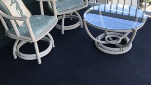 Un porche con dos sillas de mimbre blancas con cojines azules y una mesa de vidrio.
