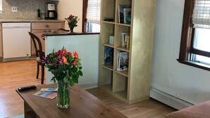 Una sala de estar con una mesa de centro de madera, un sofá, un televisor sobre un mueble bajo y una cocina al fondo.