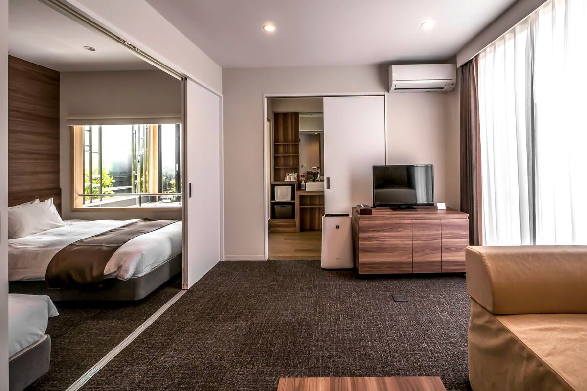 A hotel room with a bed, a sofa, a TV on a wooden cabinet, and a bathroom visible through an open door.