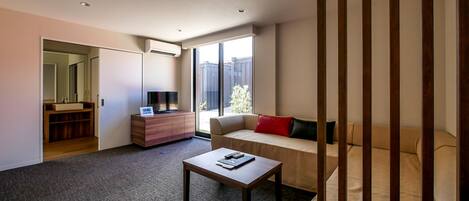 Una habitación de hotel moderna con sofá, una mesita, un televisor sobre un mueble de madera y un ventanal con vistas al exterior.