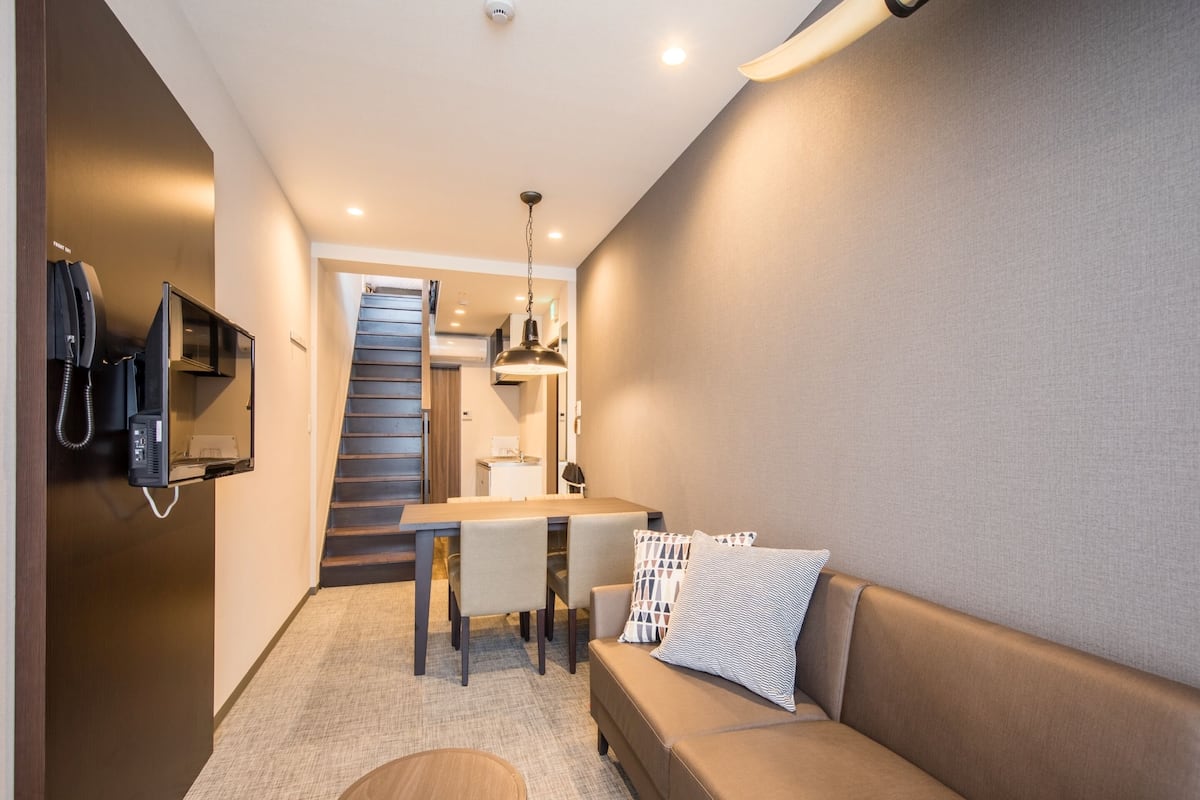 A compact hotel room with a sofa, a dining table, a small kitchen area, and a staircase.