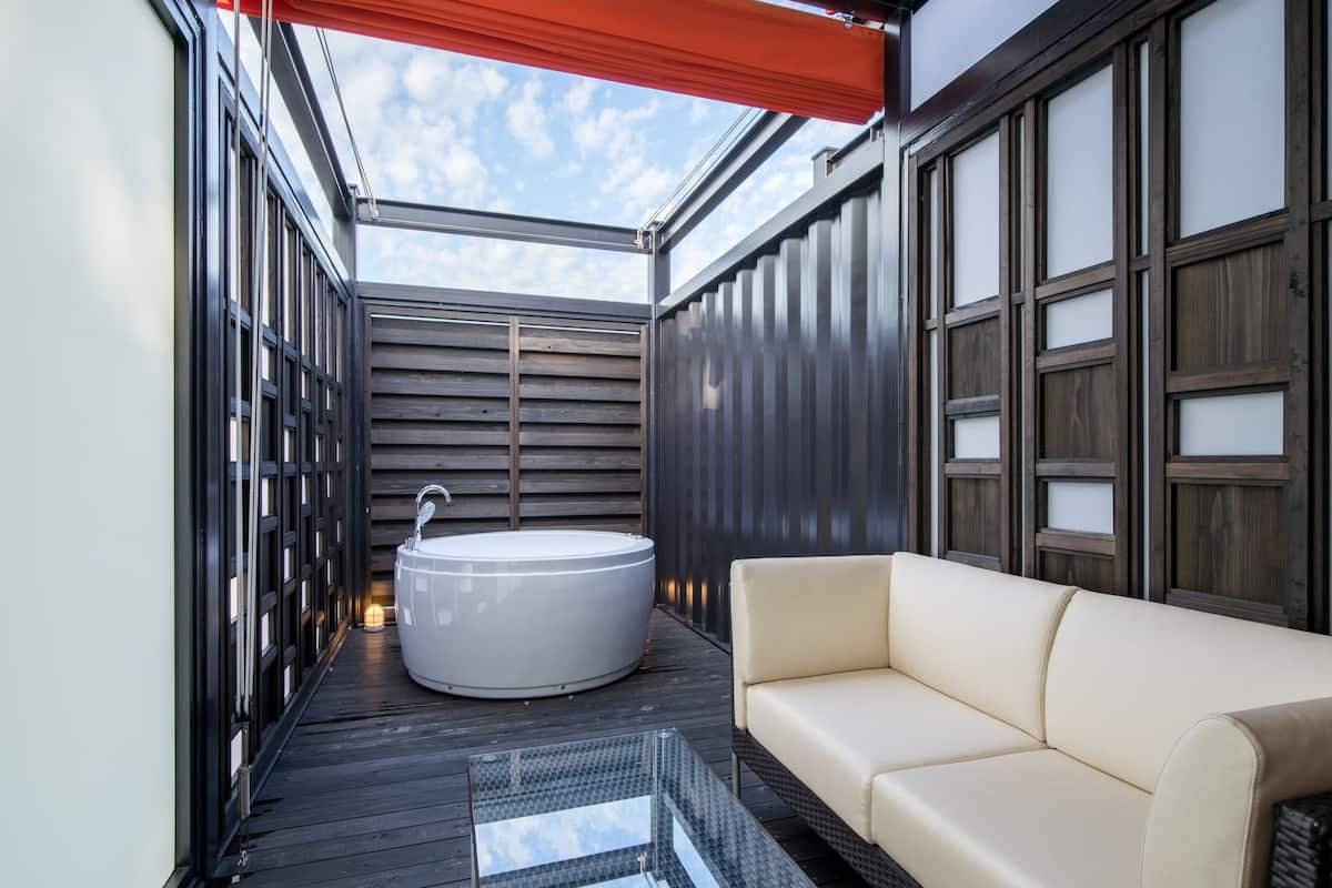 A modern outdoor bathroom with a large bathtub, a sofa, and a glass-enclosed ceiling.