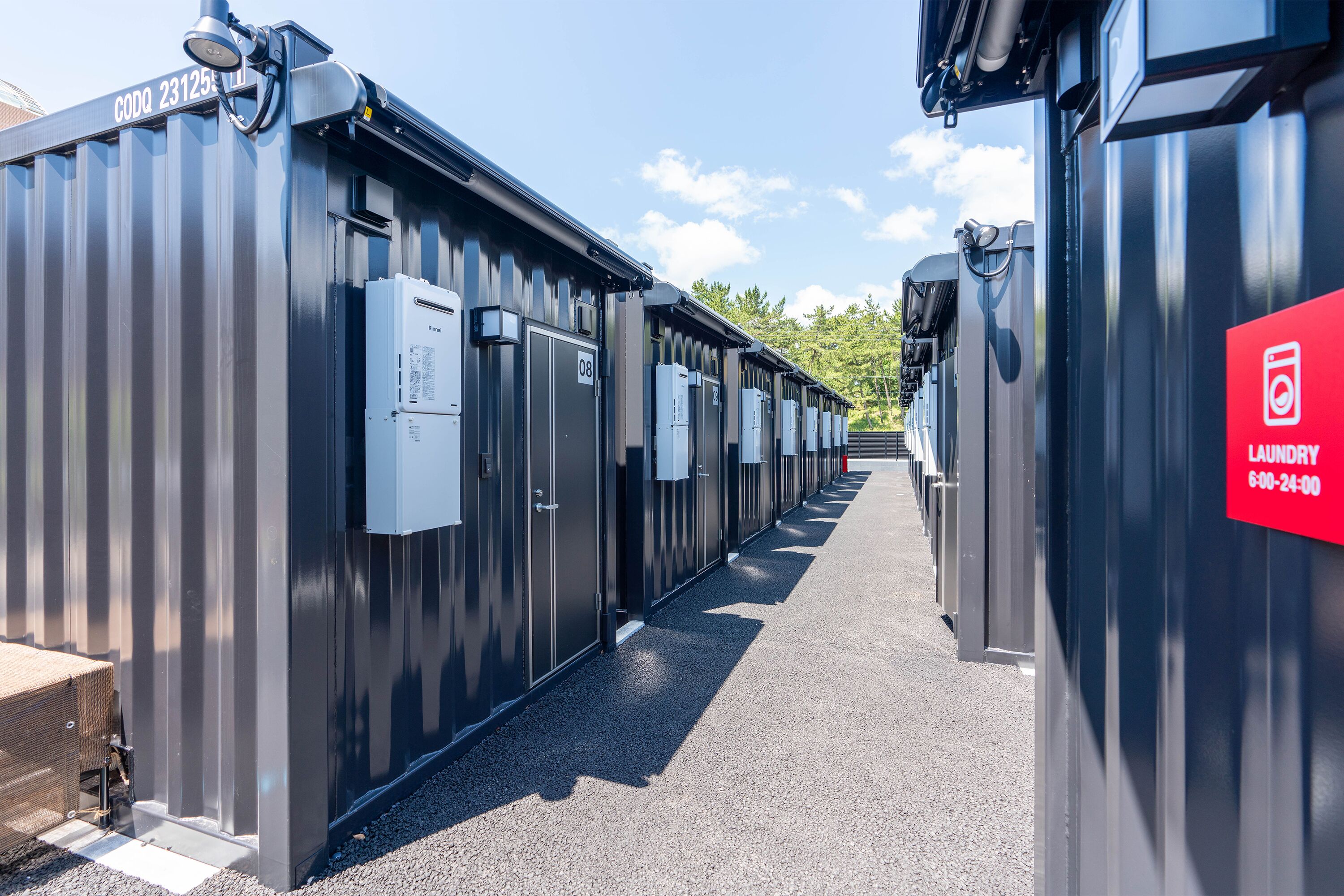 A row of industrial-style laundry facilities with numbered doors, a red sign indicating a 24-hour service, and a clear sky in the background.