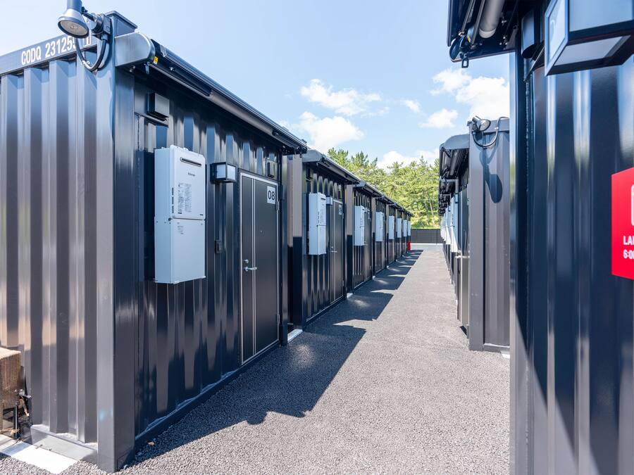 A row of industrial-style laundry facilities with numbered doors, a red sign indicating a 24-hour service, and a clear sky in the background.