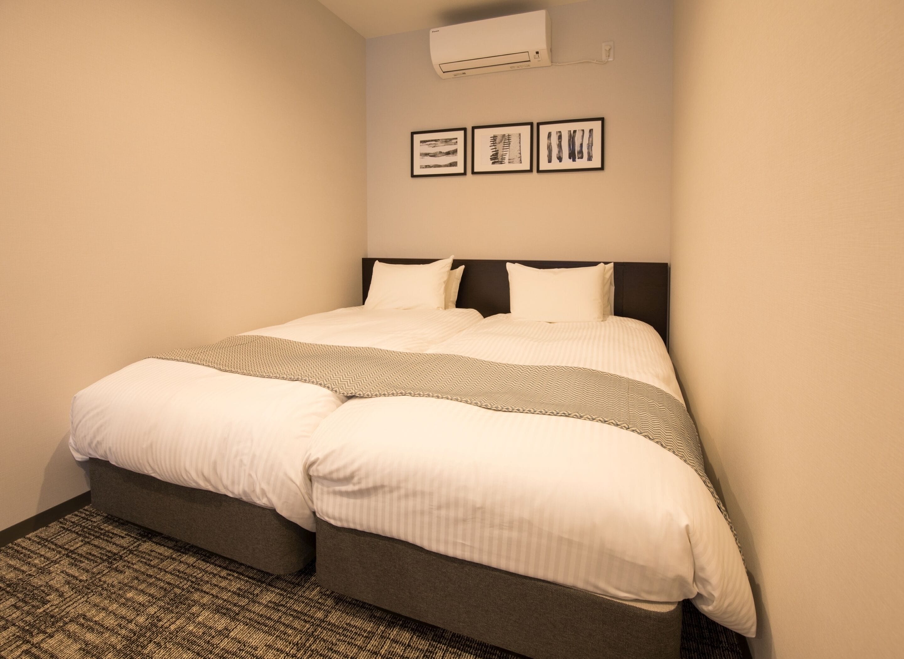 A neatly made bed with a gray headboard, a patterned throw blanket, and a plain wall with three framed pictures.