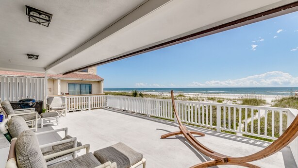 A balcony with wicker furniture and a wooden lounge chair, overlooking a beach with a clear sky. - Spacious Beachfront Condo NEWLY REMODELED (Jacksonville Beach)