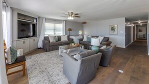 A modern living room with grey sofas, a coffee table, and a TV. There is a ceiling fan, a chandelier, and a kitchen area in the background. - Spacious Beachfront Condo NEWLY REMODELED (Jacksonville Beach)