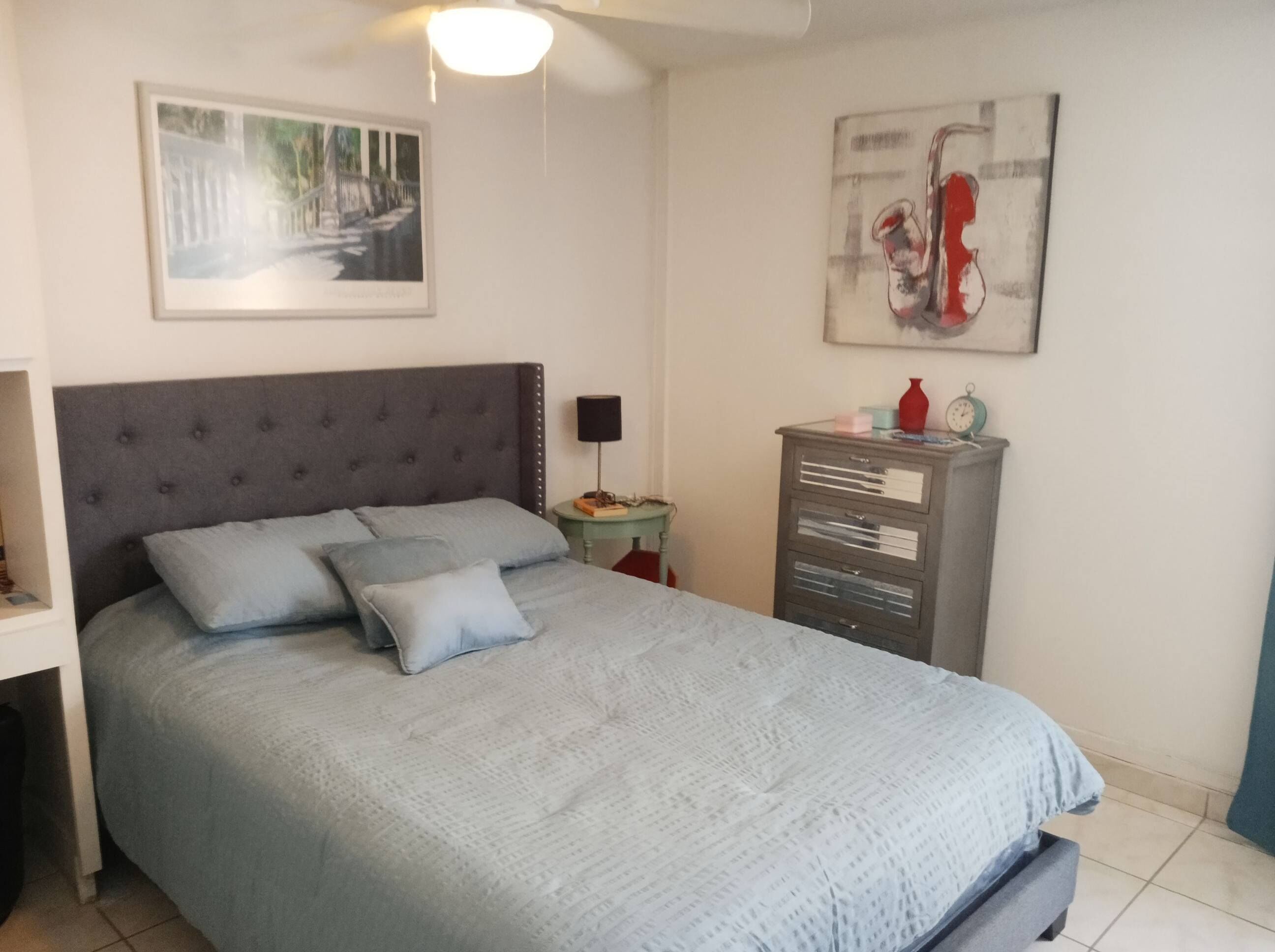 Un lit bien fait, avec une tête de lit grise, une petite table de chevet avec une lampe et une commode à tiroirs.