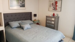 Ein ordentlich bezogenes Bett mit grauem Kopfteil, ein kleiner Nachttisch mit Lampe und eine Kommode mit Schubladen.