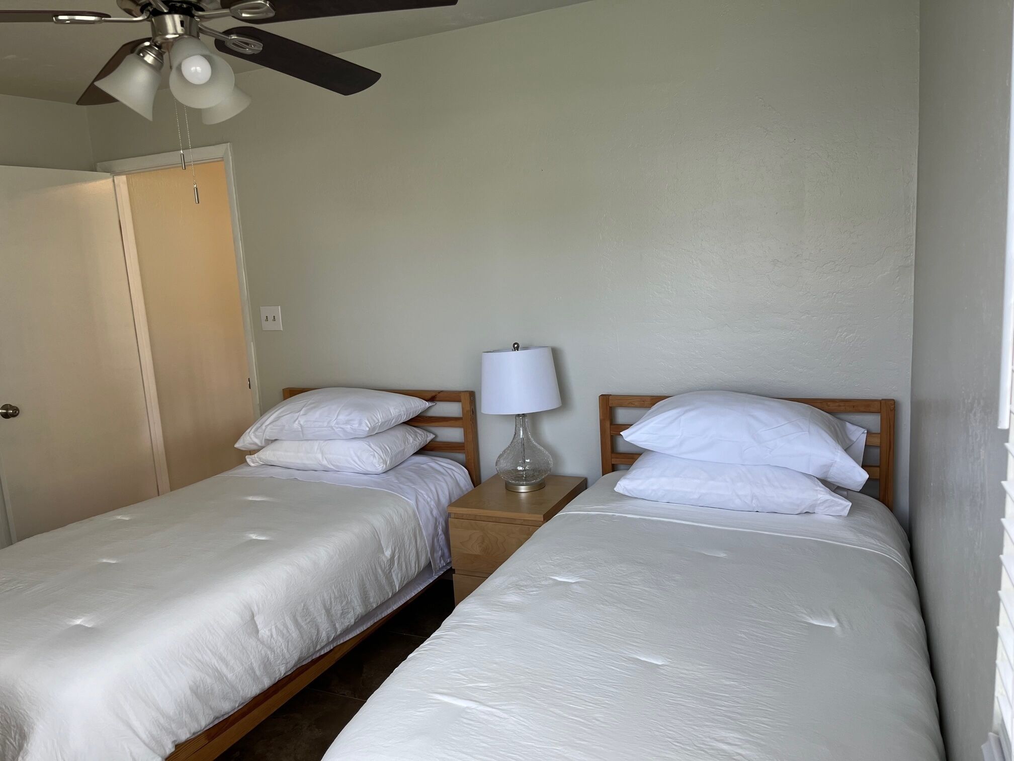 Una habitación con dos camas individuales, un ventilador de techo, una mesita de noche con lámpara y una puerta.
