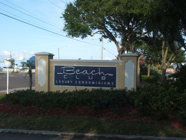 En skylt för Beach Club Luxury Condominiums, placerad så att man tydligt ser entrén och den omgivande vegetationen.