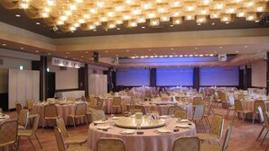 Een ruime congreszaal met ronde tafels en stoelen, een podium met een blauwe achtergrond en een plafond met een decoratieve lamp.