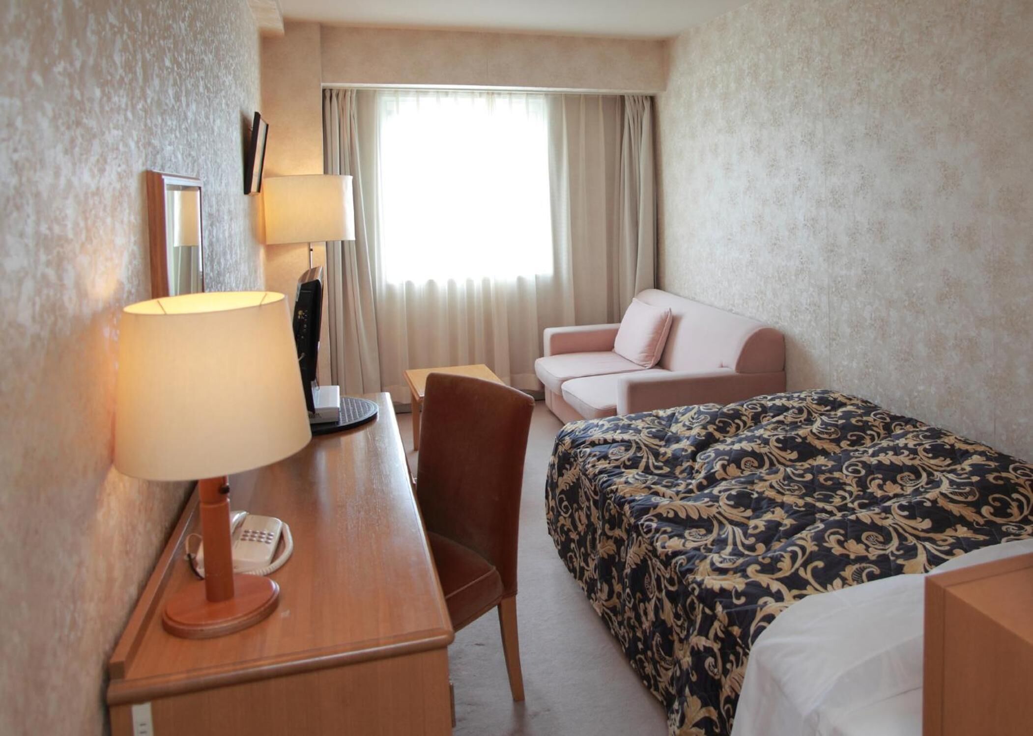 A hotel room with a bed, a desk with a lamp, a chair, a sofa, and a window with curtains.