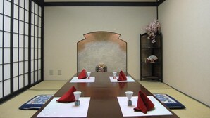 Een traditionele Japanse kamer met tatami vloerbedekking, een lage tafel gedekt met rode servetten en wit papier, en een decoratieve wand in de achtergrond.