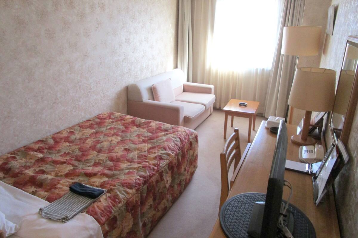 Une chambre d’hôtel comprenant un lit, un bureau avec une chaise, une petite table, une lampe et un téléviseur.