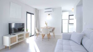 Ein modernes Wohnzimmer mit Flachbildfernseher, einem weißen Sofa, einem Essbereich mit Tisch und Stühlen sowie einer Küchenzeile mit Kühlschrank.