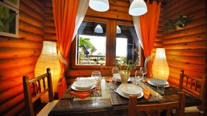 Un coin repas aménagé dans une cabane en bois, avec une table dressée pour quatre personnes, deux lampes et une fenêtre avec des rideaux.