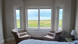 A room with a bed, two armchairs, a nightstand, and a lamp. There are three windows providing a view of the sea and grassy landscape. - Amazing Sea views.  Central location. Overlooking Portstewart Old Course. (Portstewart)