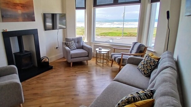 A living room with a fireplace, grey sofas, a TV, and a view of the sea. - Amazing Sea views.  Central location. Overlooking Portstewart Old Course. (Portstewart)