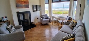 A living room with a fireplace, grey sofas, a TV, and a view of the sea. - Amazing Sea views. Central location. Overlooking Portstewart Old Course. (Portstewart)