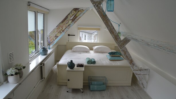 A bedroom with a sloped ceiling, a bed with white linens, a small bedside table with a kettle, and a window with a view of a house.