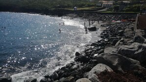 A rocky beach with clear blue water, people swimming and walking along the shore, and houses in the background.