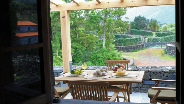 A wooden table set with food and drinks, overlooking a lush garden and stone walls.