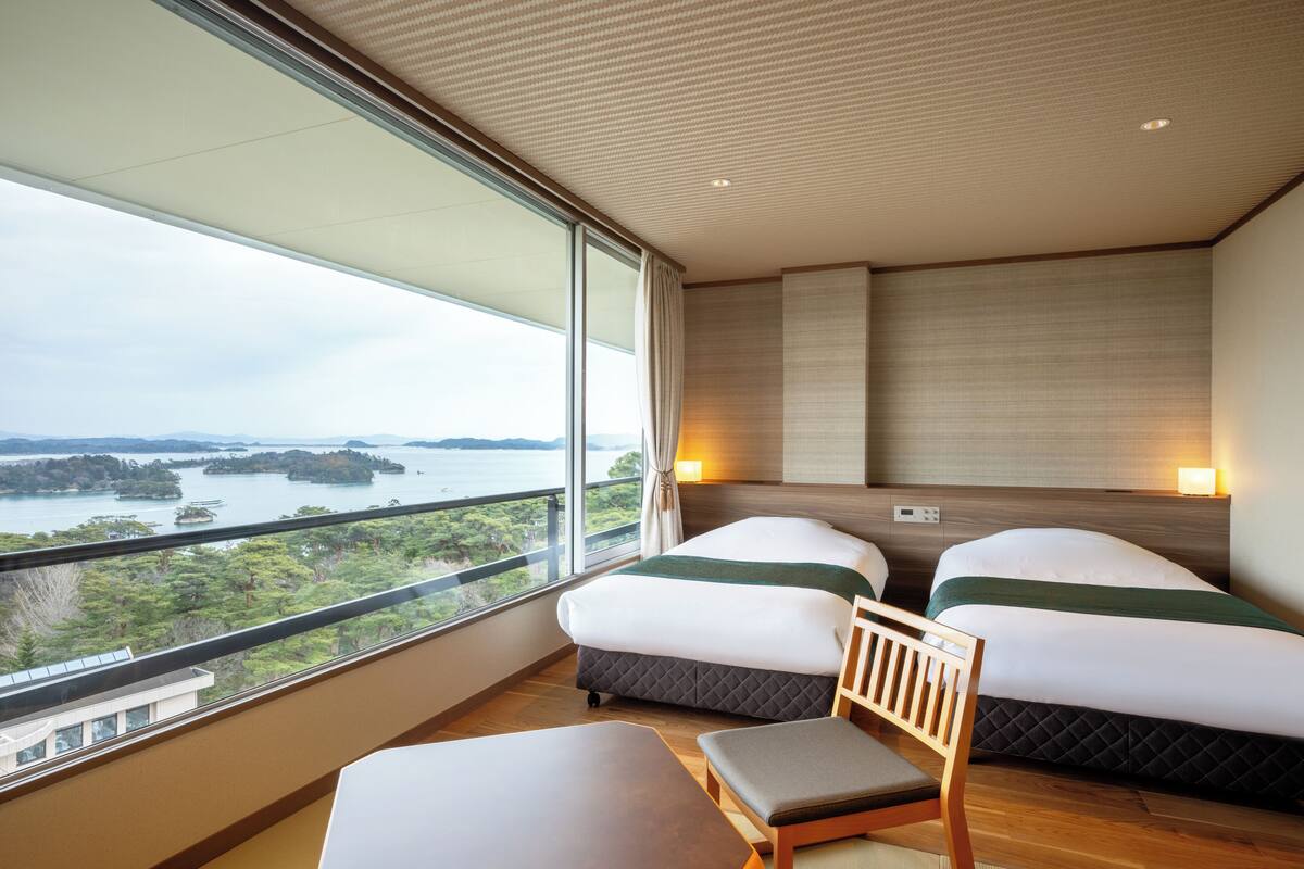 Une chambre d’hôtel avec deux lits, une vue sur la mer, un balcon et une table avec une chaise.