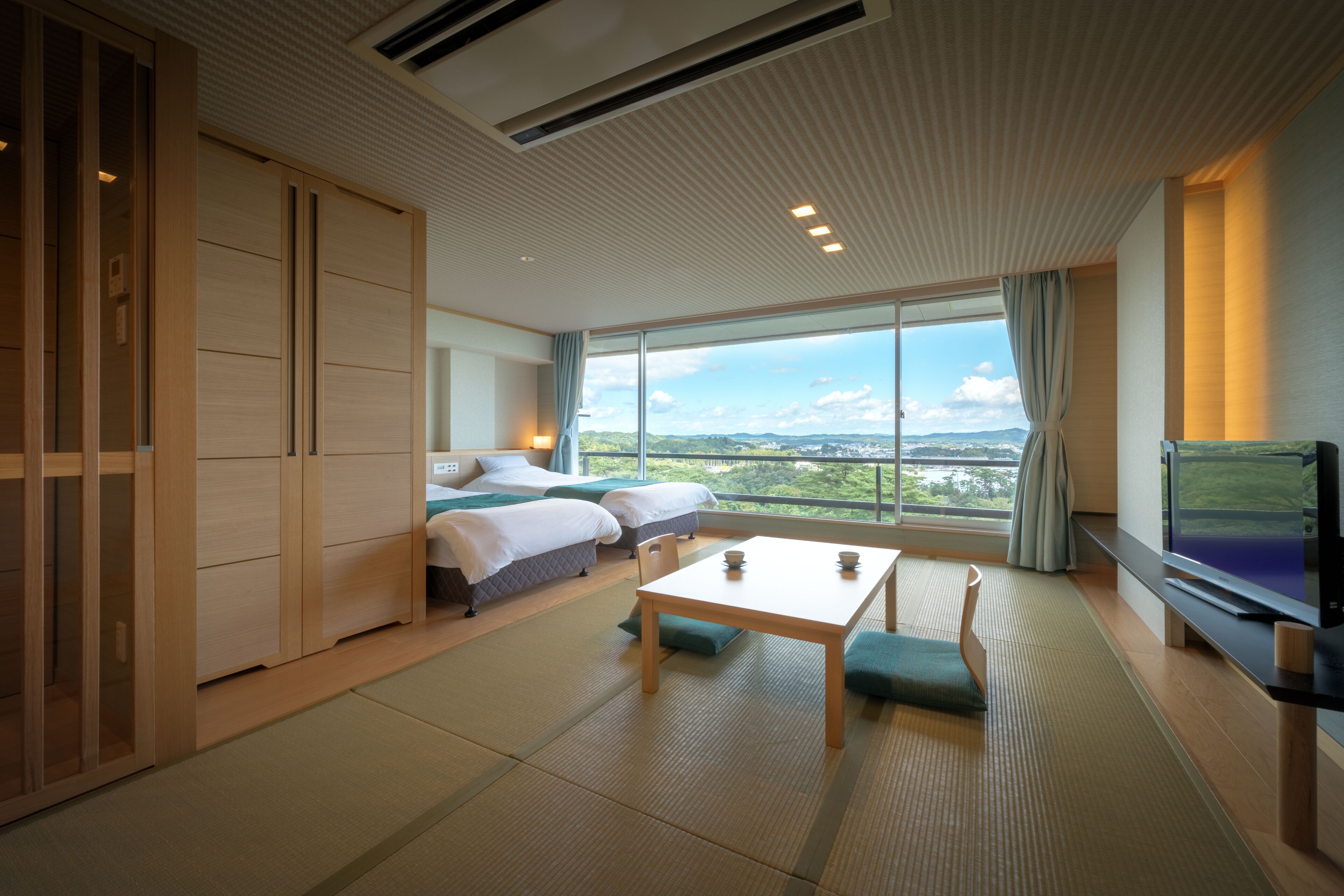 A modern hotel room with a large window, a wooden wardrobe, a bed, a table, and a television.