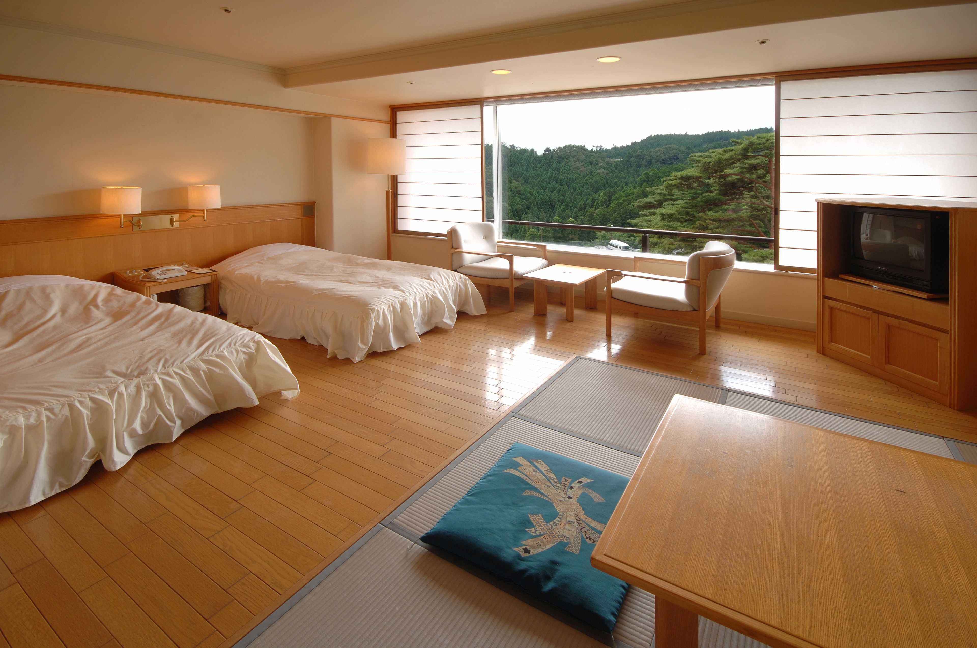 A hotel room with two beds, a TV, a table, and a view of a forested area.