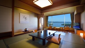 Een traditionele Japanse kamer met tatami vloerbedekking, een lage tafel met kussens, een groot raam met uitzicht op een waterpartij en een televisie aan de rechterkant.