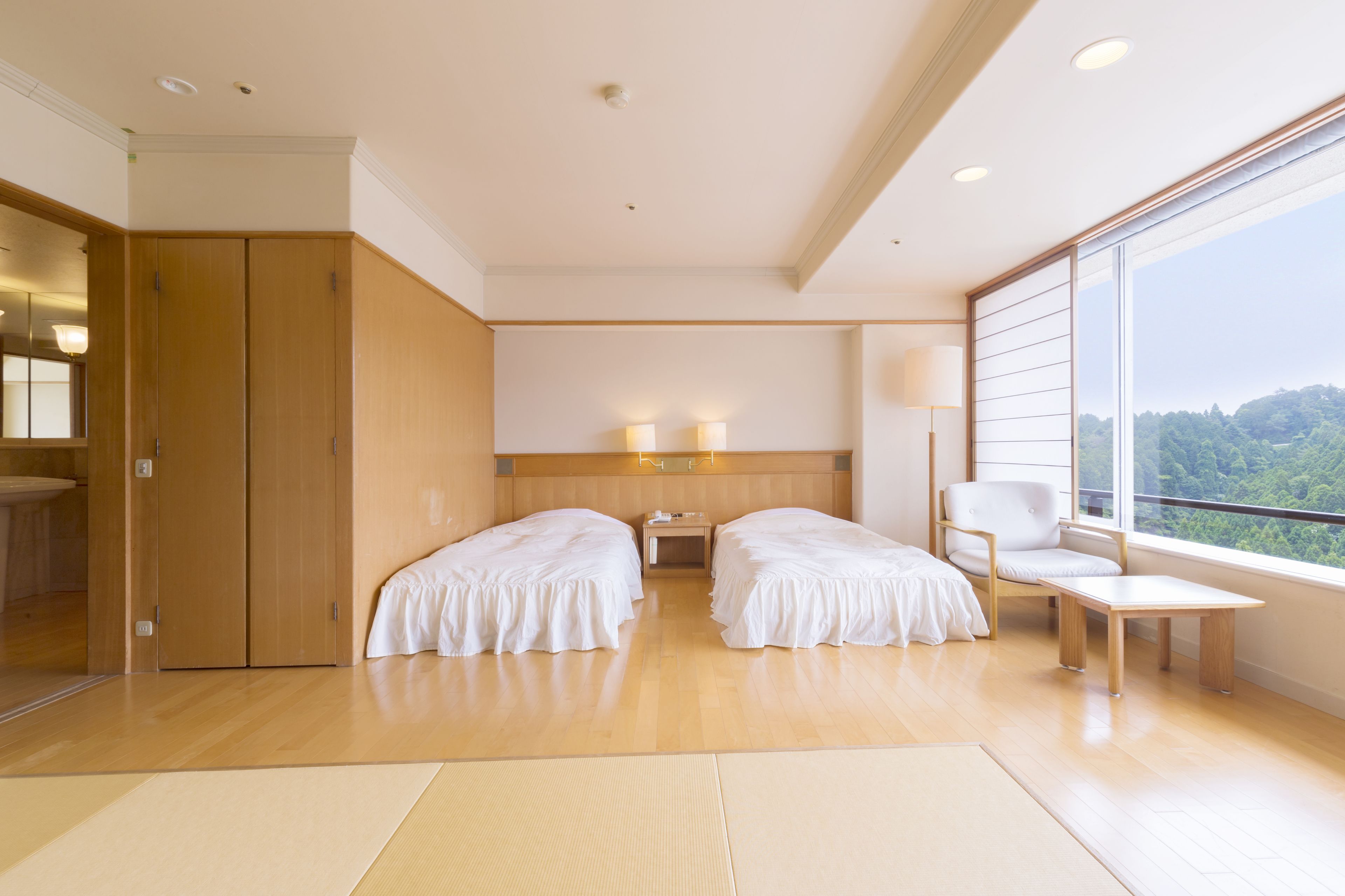 Habitación de hotel con dos camas, un armario de madera, una mesita de noche, una silla y un amplio ventanal con vistas al exterior.