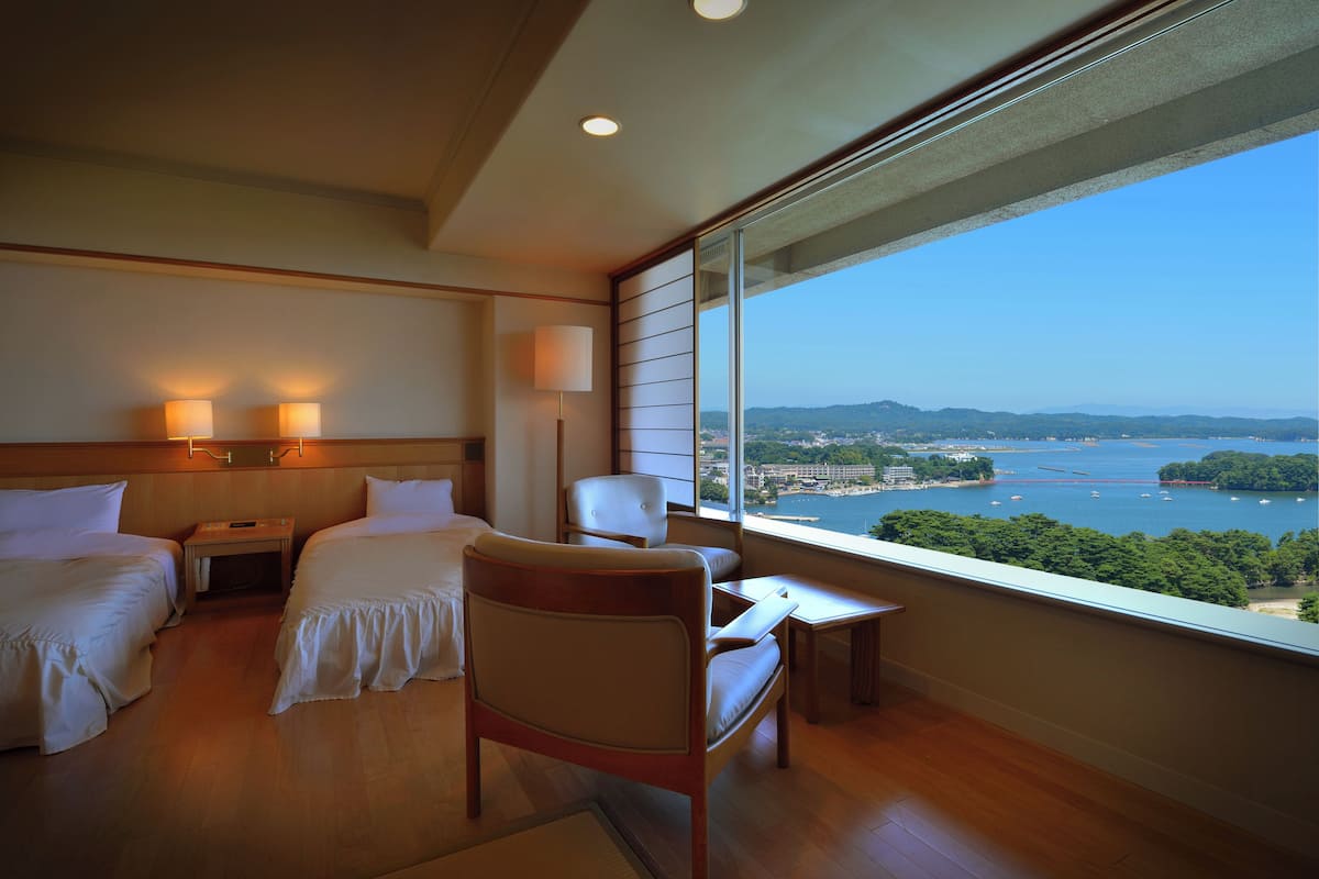 Une chambre d’hôtel avec deux lits, une chaise, une petite table et une vue sur une étendue d’eau et un paysage lointain.