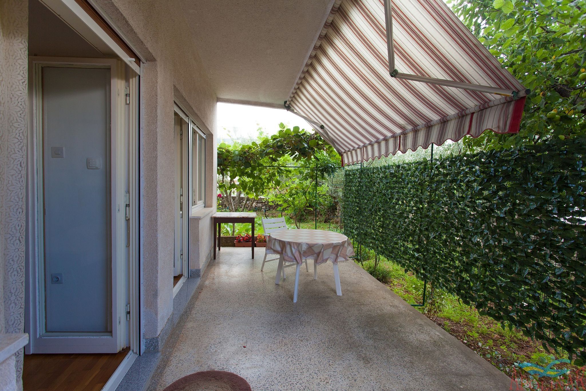 A covered patio with a table and chairs, a door, and a hedge.
