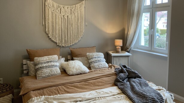 A bedroom with a bed, pillows, a macrame wall hanging, a bedside lamp, and a window with curtains. - Beautiful old building apartment in a top location 10 minutes to town in the residential area (Biederitz)