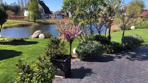 A serene garden with a pond, neatly trimmed grass, and flowering trees. - Fantastic 5 ***** Thatched cottage on the water with sauna, outdoor Jacuzzi, fireplace u. v. (Fuhlendorf)