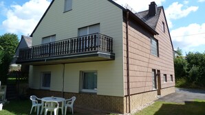 A two-story house with a balcony, a small patio with a table and chairs, and a brick foundation. - Apartment "Eifel Nest" between VULKANEIFEL and Moselle (Urschmitt)