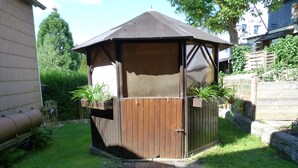A wooden gazebo with a hexagonal tiled entrance, surrounded by a grassy yard and a wooden fence. - Apartment "Eifel Nest" between VULKANEIFEL and Moselle (Urschmitt)