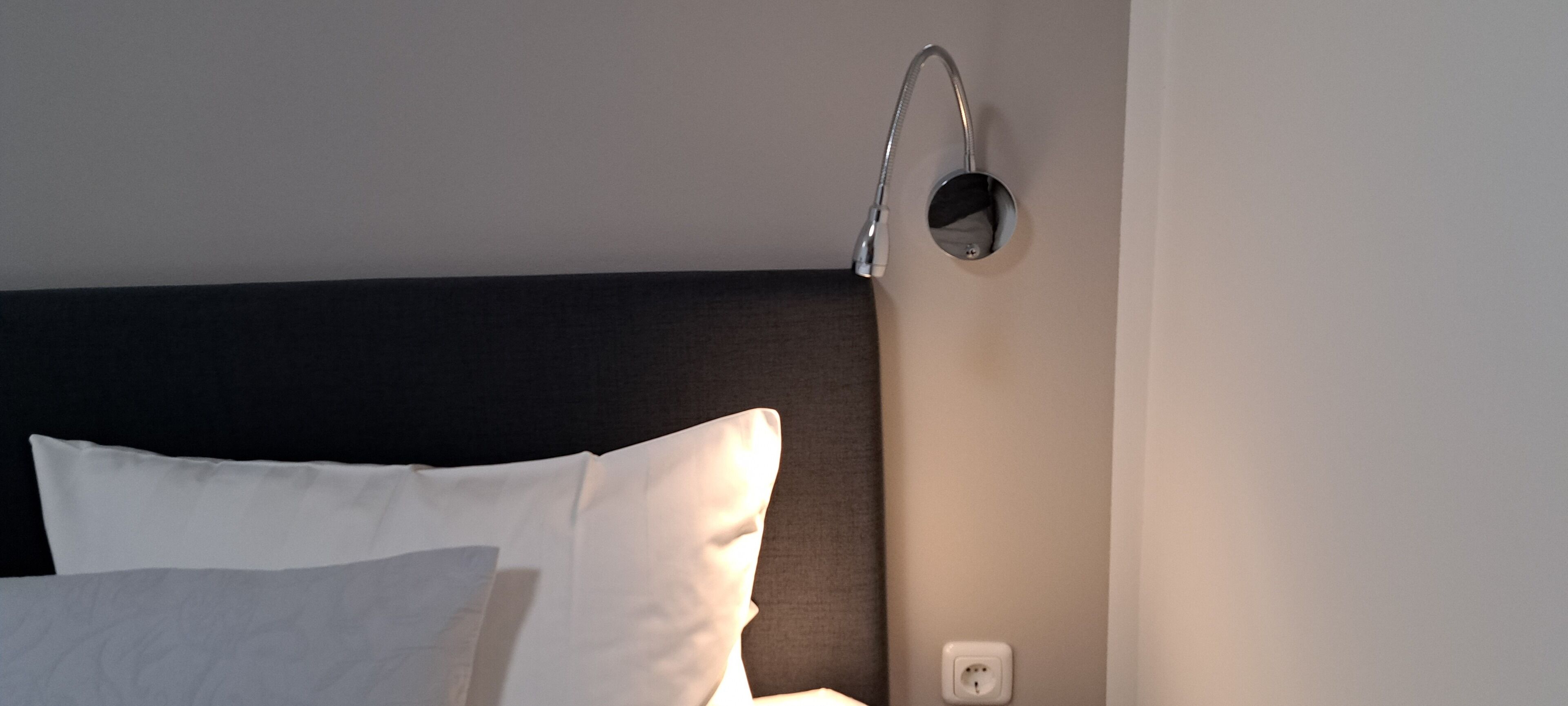 Ein ordentlich bezogenes Bett mit weißen Leinen, einem dunklen Kopfteil und einer Wandleuchte.