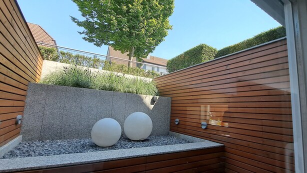 A modern outdoor seating area with wooden slatted walls, a stone bench, and two spherical white planters.