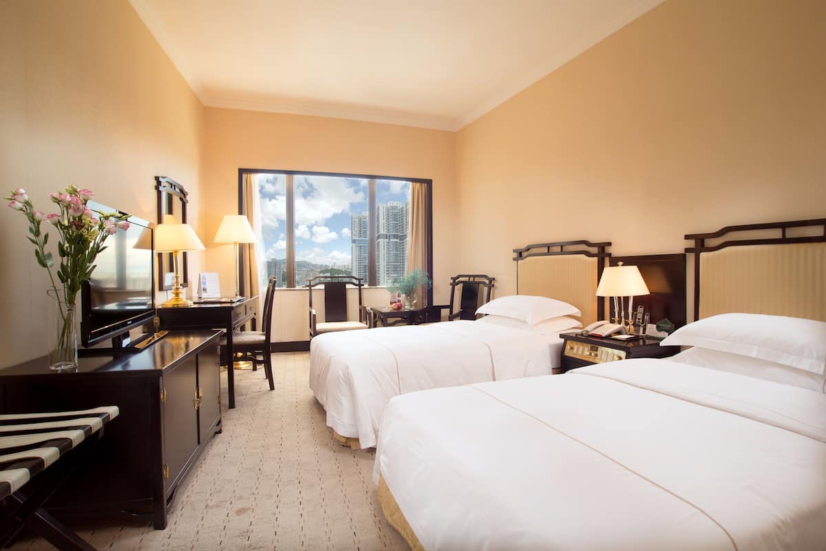 Une chambre d’hôtel avec deux lits, un bureau avec une lampe, une chaise, une commode et une grande fenêtre donnant sur la ville.
